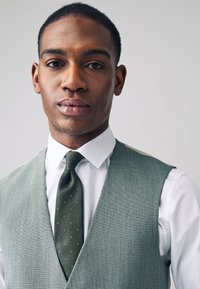 Gilet texturé gris porté sur une chemise blanche, accompagné d'une cravate vert foncé à petits pois blancs. Le design est ajusté et élégant.