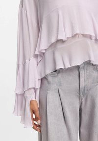 Lyse lilla, lagdelt bluse med tekstureret stof og flæsekant. Kombineret med lysegrå bukser med vide ben og folder.