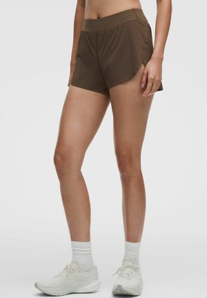 Athlete wearing brown athletic shorts, white crew socks, and white running shoes standing against a plain light background.