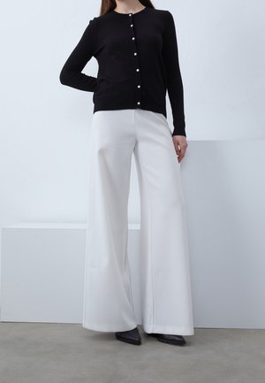 Chaqueta de punto negra con cuello redondo y botones de perla, combinada con pantalones de pierna ancha blancos. Tejido suave y corte entallado.