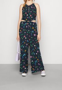 Blommönstrad jumpsuit med marinblå bakgrund, halterneck, knytmidja och vida byxor. Kompletteras med en ljuslila handväska.
