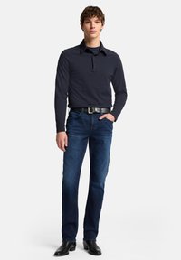 Polo a maniche lunghe blu navy abbinata a jeans in denim scuro. Cintura nera con fibbia argentata. Modello in piedi in un ambiente neutro.