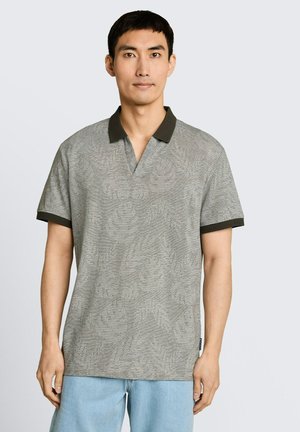 Homme portant un polo gris à manches courtes avec motifs, col et poignets noirs, assorti à un jean bleu clair, debout devant un fond uni.