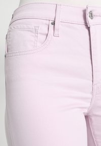Primer plano de unos pantalones de mezclilla rosa claro que muestra el bolsillo delantero, el lazo del cinturón, las costuras y el detalle del botón plateado sobre un fondo blanco.