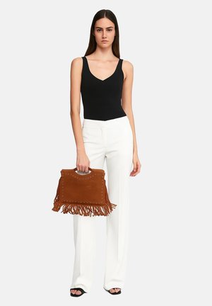 Borsa in suede tan con frange e dettagli in metallo, tenuta da una persona che indossa un top senza maniche nero e pantaloni bianchi.