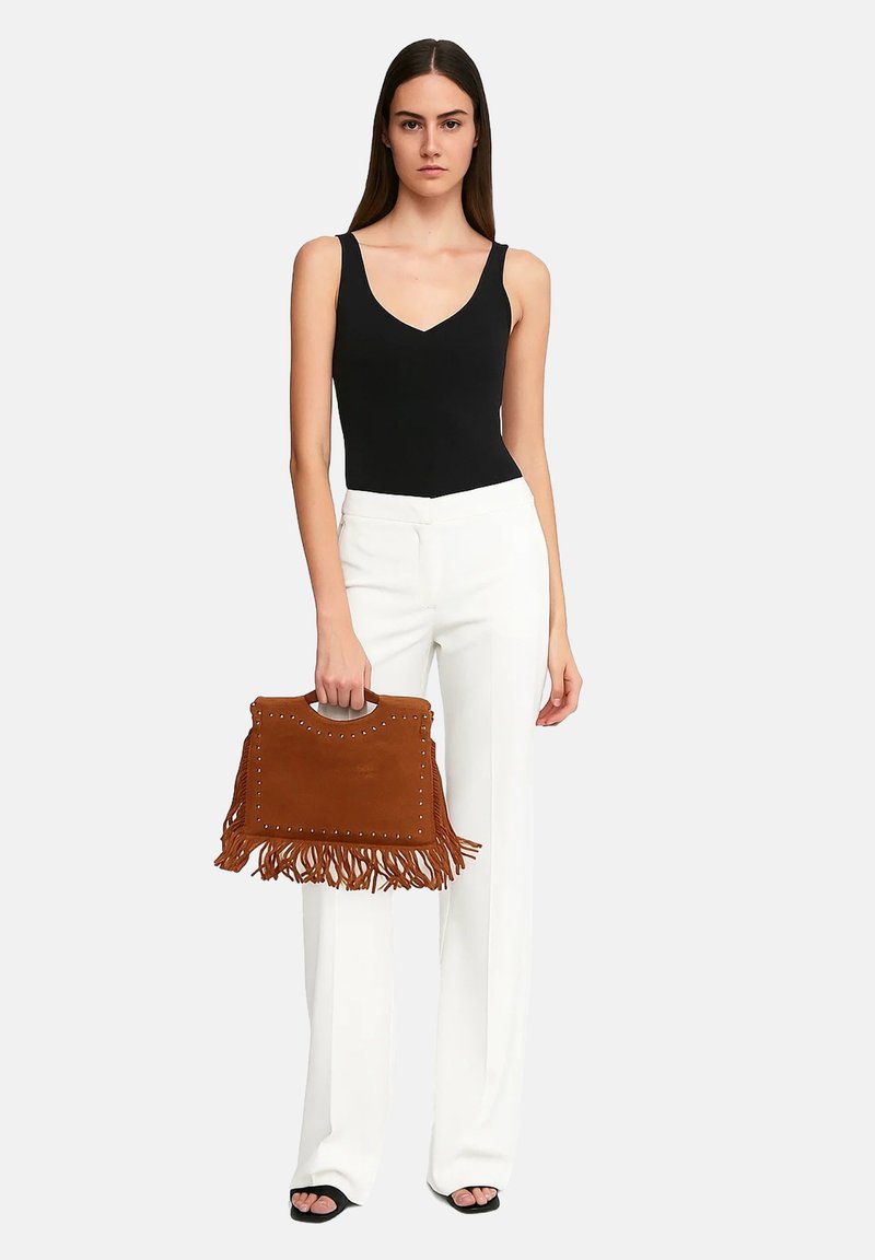 Borsa in suede tan con frange e dettagli in metallo, tenuta da una persona che indossa un top senza maniche nero e pantaloni bianchi.