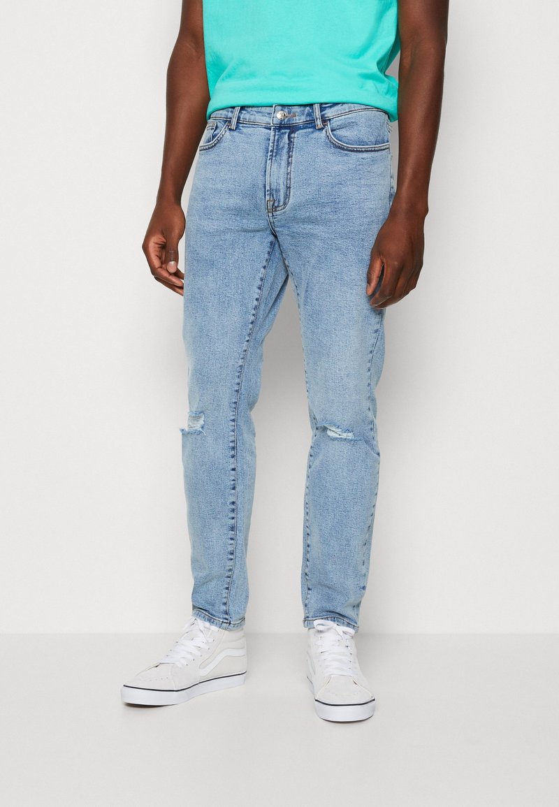 Ljust blå denimjeans med slim fit, med revor vid knäna, bärs tillsammans med vita sneakers. T-shirten är turkos och har rund halsringning.