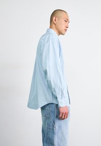 Lichtblauwe button-up shirt met verticale witte strepen, lange mouwen en een relaxte pasvorm, gedragen over blauwe jeans met een middenzak.