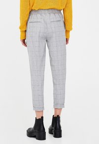 Pantalons à carreaux gris avec taille élastique, longueur cheville, deux poches arrière, associés à des bottines noires sur fond blanc.