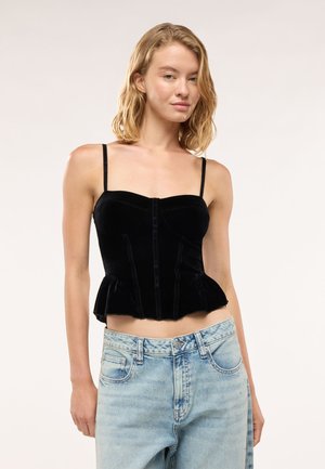 Giovane donna che indossa un top senza maniche in velluto nero con sottili spalline e jeans azzurri a vita alta, in piedi contro uno sfondo semplice.