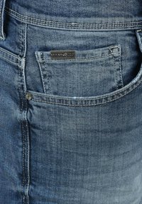 Jeans de mezclilla azul con un pequeño bolsillo frontal y detalles cosidos. Se puede ver una etiqueta de metal de la marca encima del bolsillo.