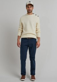 Cremiger Strickpullover mit Textur und Knöpfen an den Schultern, kombiniert mit dunkelblauen Hosen und braunen Schnürschuhen. Die Mütze hat ein Logo und einen kontrastierenden Schirm.