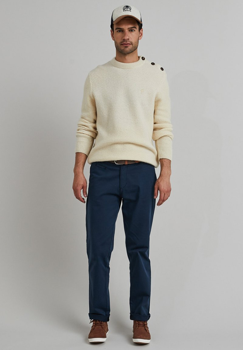 Cremiger Strickpullover mit Textur und Knöpfen an den Schultern, kombiniert mit dunkelblauen Hosen und braunen Schnürschuhen. Die Mütze hat ein Logo und einen kontrastierenden Schirm.