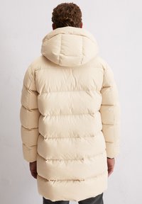 Crèmekleurig gewatteerd jack met capuchon, voorzien van een doorgestikt ontwerp, afgeronde schouders en elastische manchetten. Het materiaal lijkt zacht en licht van gewicht.