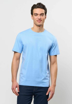 Joven con cabello oscuro que lleva una camiseta de manga corta azul claro lisa y vaqueros azul oscuro, posando frente a un fondo blanco.