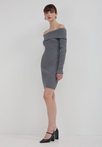 Graues, geripptes Off-Shoulder-Kleid mit langen Ärmeln, figurbetonter Silhouette und mittellanger Länge bis zur Mitte des Oberschenkels. Getragen mit schwarzen High Heels.