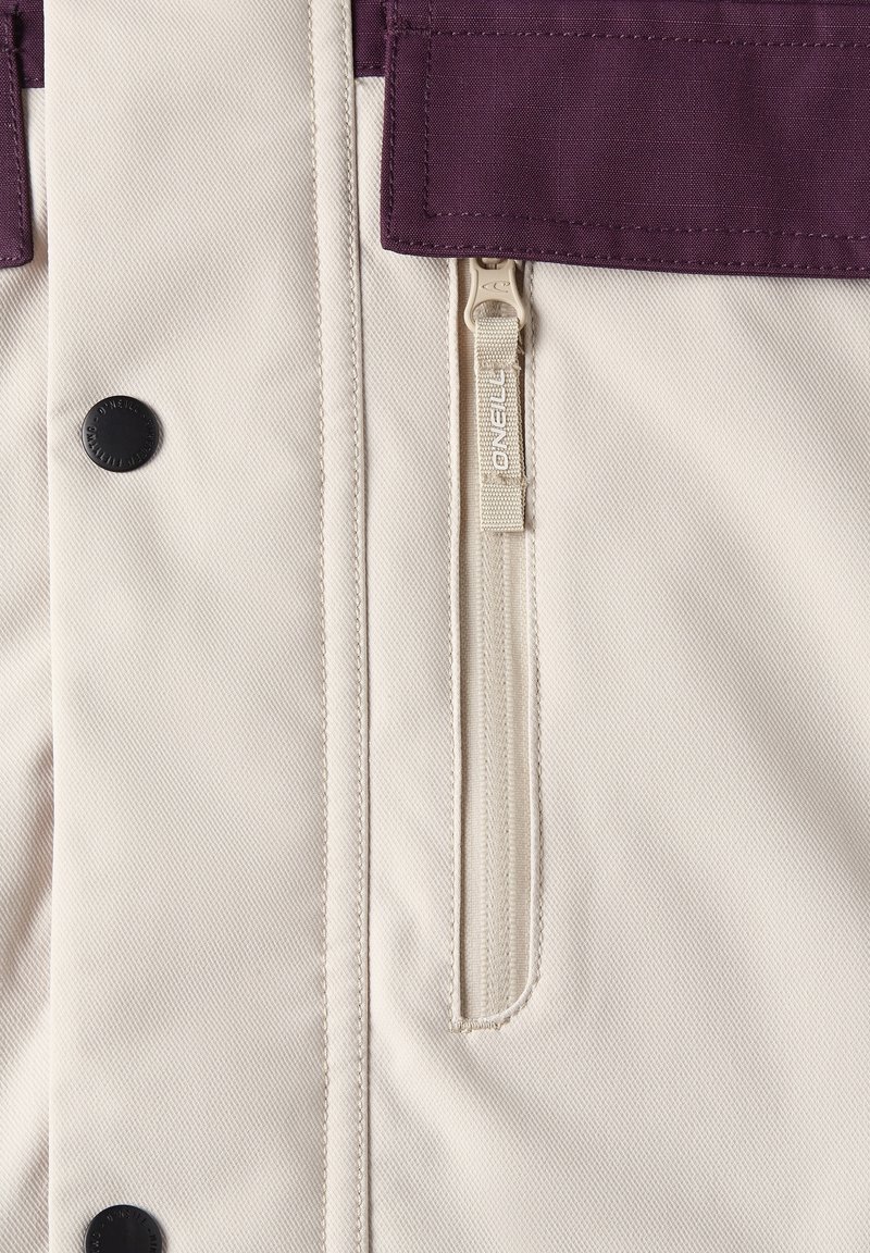 Beżowa tkaninowa kurtka z fioletowym panelem u góry, z zamkiem błyskawicznym i czarnymi elementami metalowymi. Teksturowany, wytrzymały materiał.