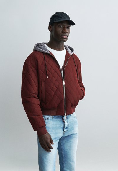 Homme portant une veste matelassée rouge foncé avec une capuche grise, une chemise blanche, un jean bleu clair et une casquette noire, debout devant un fond gris uni.