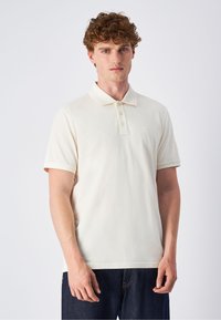 Cremfarbener Poloshirt mit klassischem Kragen, drei-Knopf-Leiste, kurzen Ärmeln und dezentem Logo-Akzent auf der Brust. Glatte Baumwolltextur.