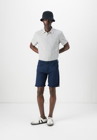 Chemise à manches courtes à rayures beige clair et marine, accompagnée d'un short bleu foncé. Accentuée par des ourlets retroussés et des baskets blanches.