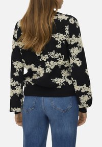 Pull noir avec motif floral crème, manches longues et ourlet côtelé. Porté avec un jean bleu. Texture douce, coupe décontractée.