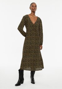 Zwarte jurk met lange mouwen en V-hals, voorzien van een gele bloemenpatroon. Midi-lengte met een vloeiende silhouet, gecombineerd met zwarte enkellaarzen.