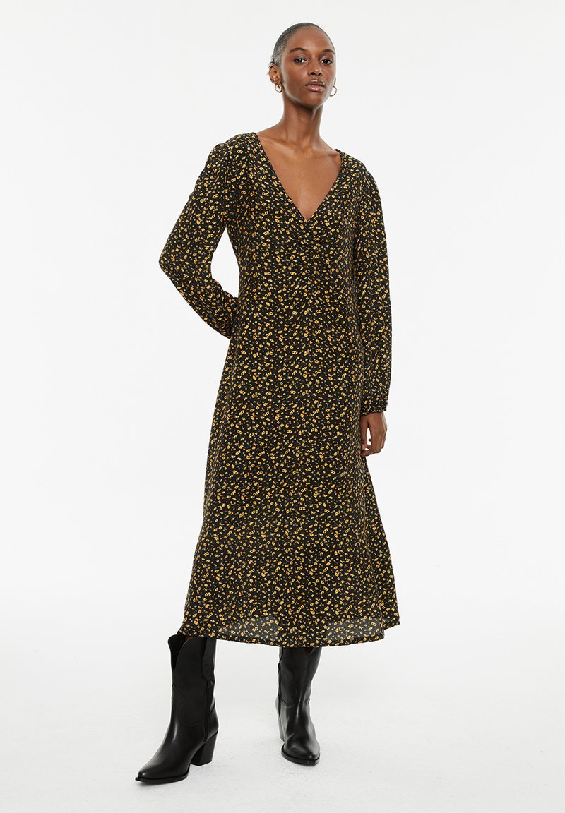 Zwarte jurk met lange mouwen en V-hals, voorzien van een gele bloemenpatroon. Midi-lengte met een vloeiende silhouet, gecombineerd met zwarte enkellaarzen.