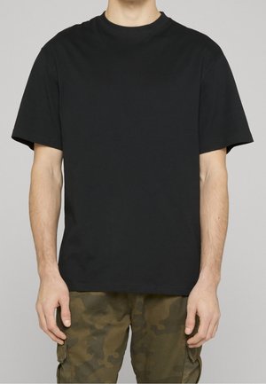 Mann trägt ein schlichtes schwarzes T-Shirt mit kurzen Ärmeln und grüne camouflage Cargohosen, steht vor einem neutralen grauen Hintergrund.