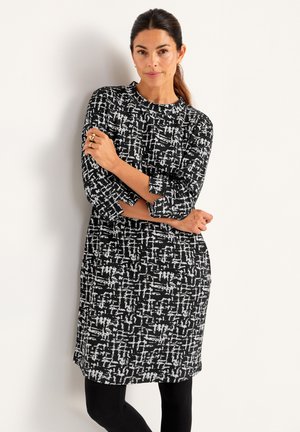 Kvinna som står mot en vit vägg, klädd i en svart- och vitmönstrad knälång klänning med trekvartsärmar och svarta leggings.