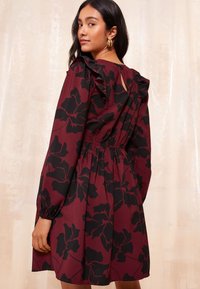 Sieviete valkā tumši sarkanu kleitu ar lielu melnu ziedu rakstu, garām pūstām piedurknēm, volāniem plecu daļā un atklātu muguras daļu ar atslēgas caurumu, skatoties pār plecu.