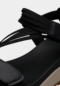 Sandale noire avec plusieurs fines lanières élastiques et une semelle épaisse et rembourrée, montrant des coutures détaillées et la texture sur la lanière et la semelle intérieure.