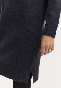 Stickad klänning i mörk marinblå med en strukturerad, mjuk yta. Inkluderar sidofickor och ribbade manschetter. Hand som vilar på klänningen.