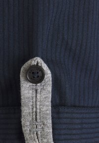 Námořnický pruhovaný materiál s texturovaným šedým pleteným popruhem, se černým knoflíkem označeným "s.Oliver." Detail knoflíku na rozhraní materiálů.