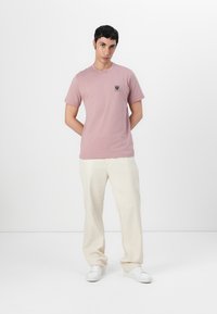 Tricou din bumbac roz deschis, cu guler rotund și mâneci scurte, având o mică aplicație grafică pe pieptul stâng, asortat cu pantaloni albaștri.