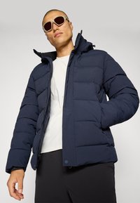 Marineblå puffjakke med glidelås foran, polstret design og justerbar hette. Har sidelommer og et jevnt, teksturert stoff.