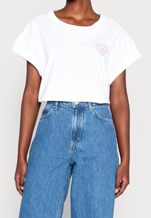 White cotton t-shirt with rolled sleeves and a small pink heart graphic on the left chest, paired with high-waisted blue denim jeans.