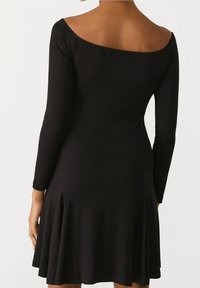 Musta mekko, jossa on pitkät hihat, olkaimeton kaula-aukko, liehukas helma, valmistettu pehmeästä kankaasta, jossa on hienovaraisia poimutuksia helmassa.