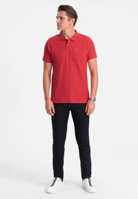 Polo rouge à col court, associé à un jean sombre et des baskets blanches. Présente un logo discret sur la poitrine.