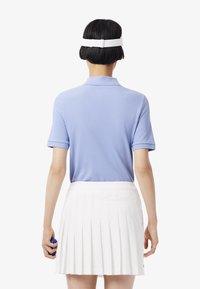 Camisa polo corta azul claro con cuello, combinada con una falda blanca con pliegues. Diadema blanca visible en la parte de atrás.