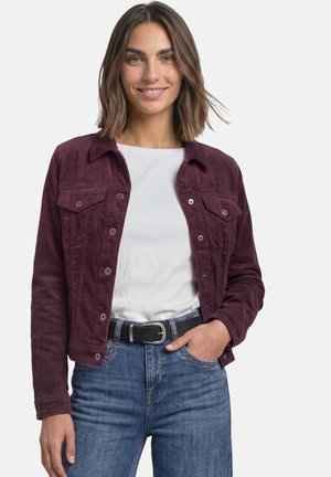Femme aux cheveux bruns mi-longs, portant une veste bordeaux, une chemise blanche, un jean bleu et une ceinture noire, souriante avec une main dans la poche.