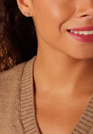 Boucles d'oreilles en forme de cœur ton doré avec un accent en gemstone claire, portées par une personne aux cheveux bouclés et portant un pull en tricot beige.