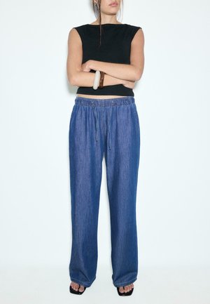 Žena so založenými rukami, oblečená v čiernom tielku bez rukávov, modré voľné nohavice so šnúrkou, sandále a hrubé náramky na jednom zápästí.