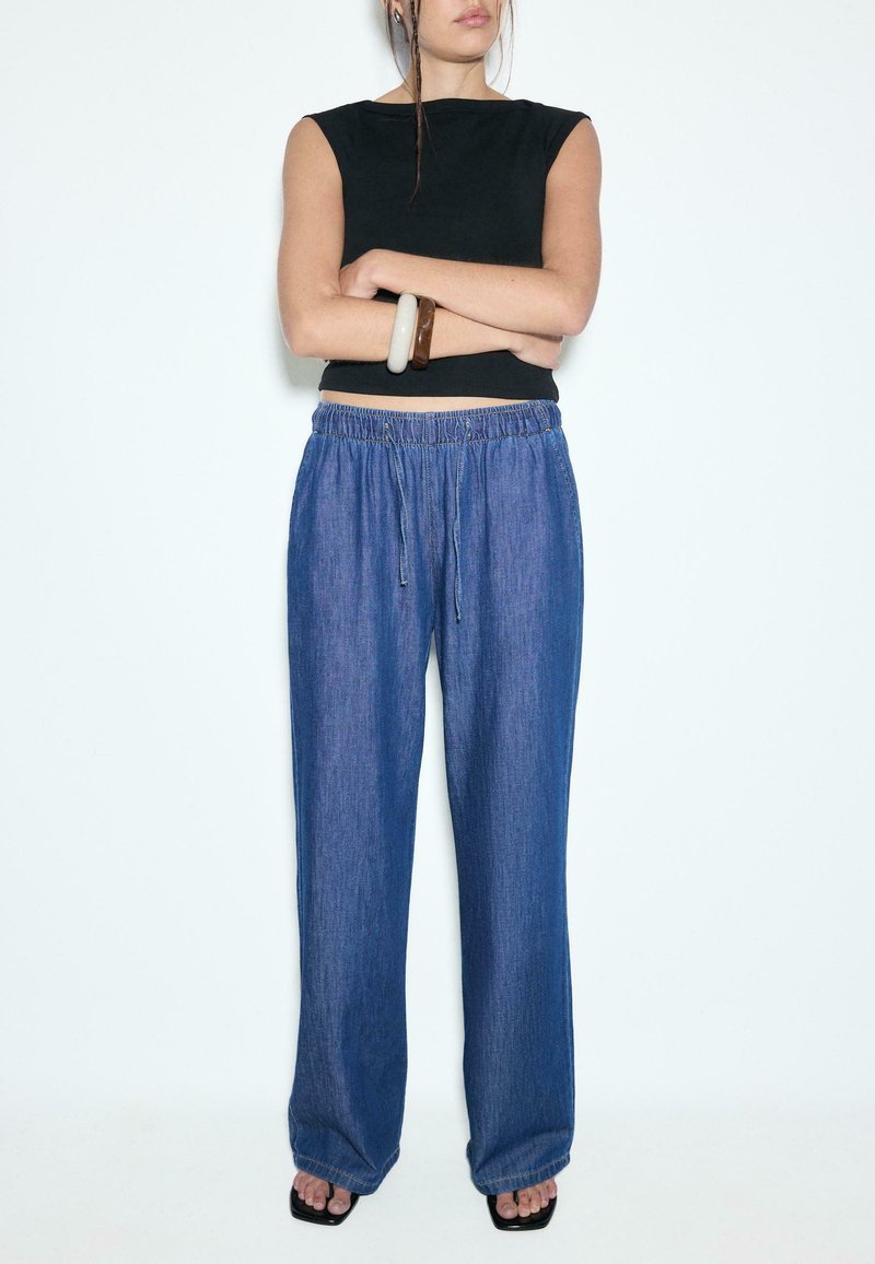 Femme les bras croisés portant un top noir sans manches, un pantalon bleu ample avec cordon, des sandales et des bracelets volumineux à un poignet.