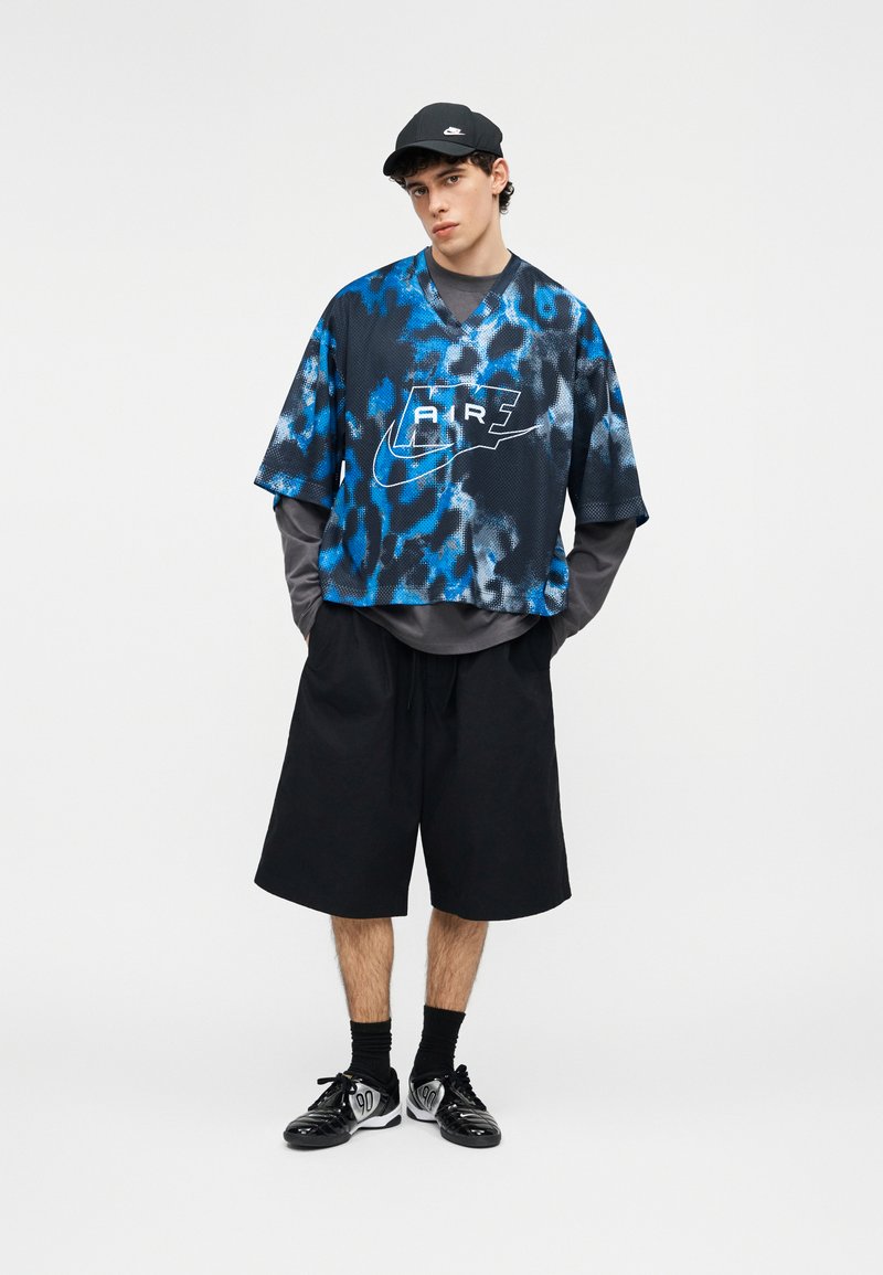 Junger Mann mit schwarzer Nike-Kappe, blau-schwarz gemustertem Nike Air Trikot über grauem Langarmshirt, schwarzen weiten Shorts, Socken und Sneakers.