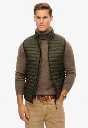 Groene puffer vest met horizontale stiksels, voorrits en hoge kraag, gedragen over een lichtbruin geribbeld sweater en beige broek.