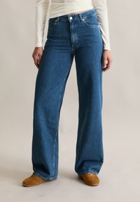 Osoba ubrana w niebieskie, szerokie dżinsy z wysokim stanem, białą, długą bluzkę i brązowe, zamszowe buty, stojąca na neutralnym tle.