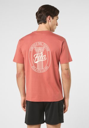 Uomo con maglietta corallo con logo vintage FILA da tennis bianco e pantaloncini neri, in piedi di spalle su uno sfondo semplice.