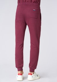 Pantaloni sportivi color bordeaux con cintura elastica, caviglie affusolate e una tasca posteriore con un piccolo logo tricolore. Materiale in cotone morbido.
