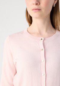 Vaaleanpunainen neuletakki, jossa on pyöreä pääntie, edessä nappisuljenta kultasävyisillä napeilla sekä pehmeä ja sileä neuletekstuuri.