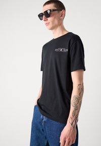 Black cotton t-shirt with short sleeves, featuring a graphic logo print. Worn with dark blue denim jeans and large black sunglasses.